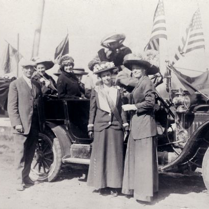 A group of women and one man stand around a car and the US Flag in a black and white photos